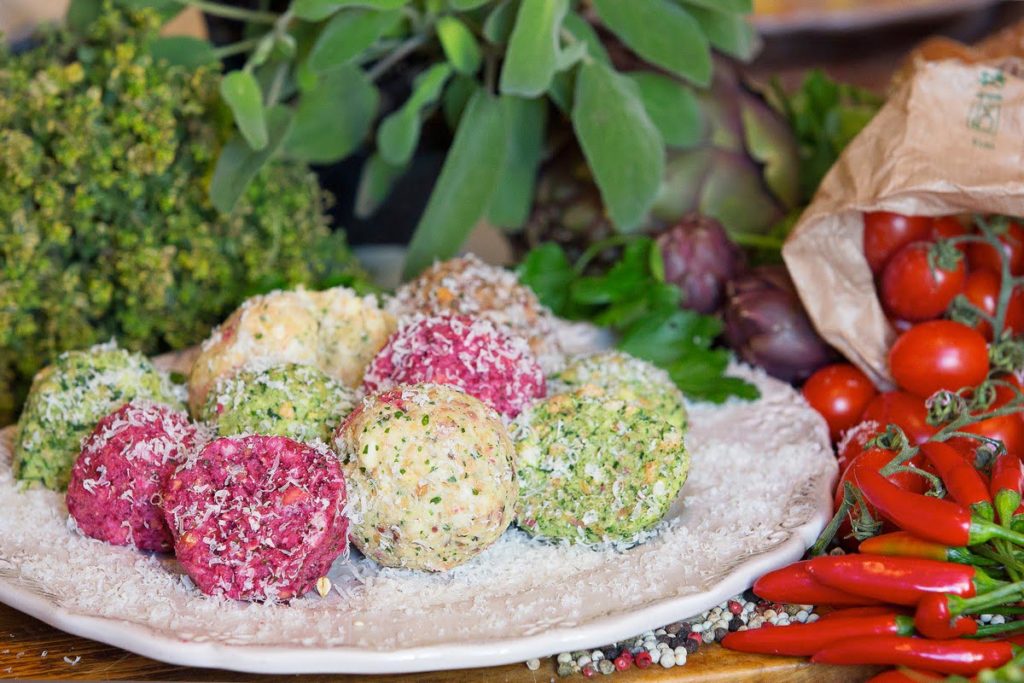 10 Piatto di polpette colorate preparate con ingredienti freschi, tra cui erbette e spezie, disposte su un piatto bianco con un leggero strato di formaggio grattugiato, circondate da pomodorini, peperoncini e verdure fresche sullo sfondo.