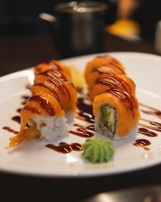 fooda_012 Rotoli di sushi con salmone fresco, avocado e tempura, guarniti con salsa scura e serviti su un piatto bianco con una porzione di wasabi in primo piano, fotografia professionale con sfondo sfocato per mettere in risalto il dettaglio del piatto.