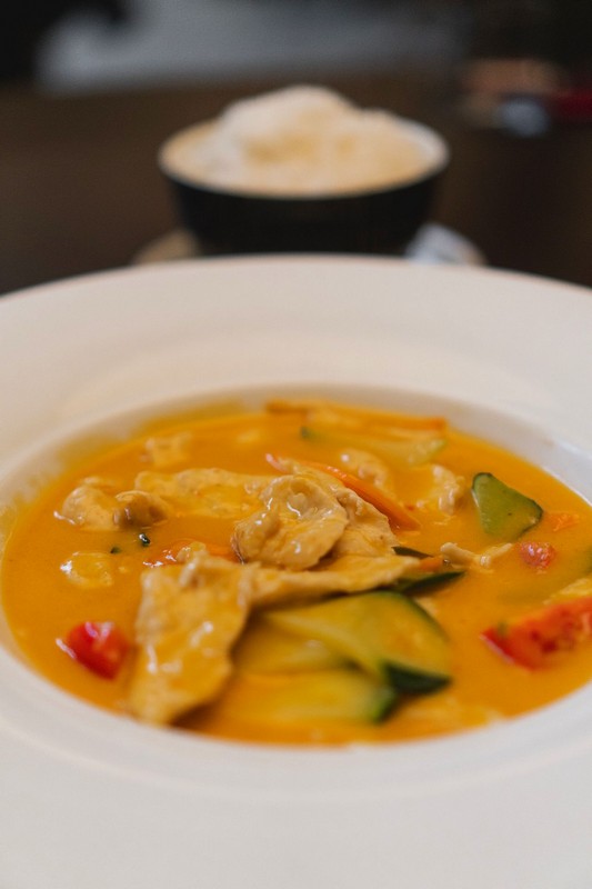 fooda_013 Zuppa cremosa con pezzi di carne e verdure fresche come zucchine e peperoni, servita in una ciotola bianca con un piatto di riso sullo sfondo, con sfocatura artistica che evidenzia il piatto principale.