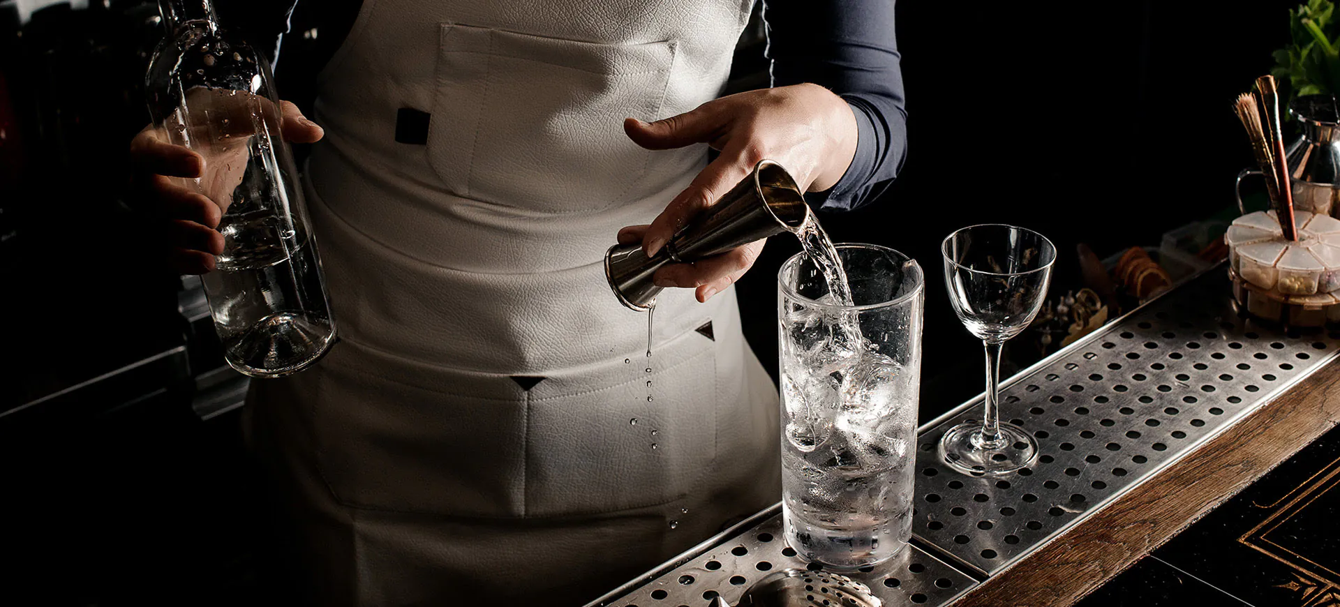 Barman con grembiule bianco che versa un distillato trasparente con un jigger in un bicchiere pieno di ghiaccio sul bancone di un bar elegante.