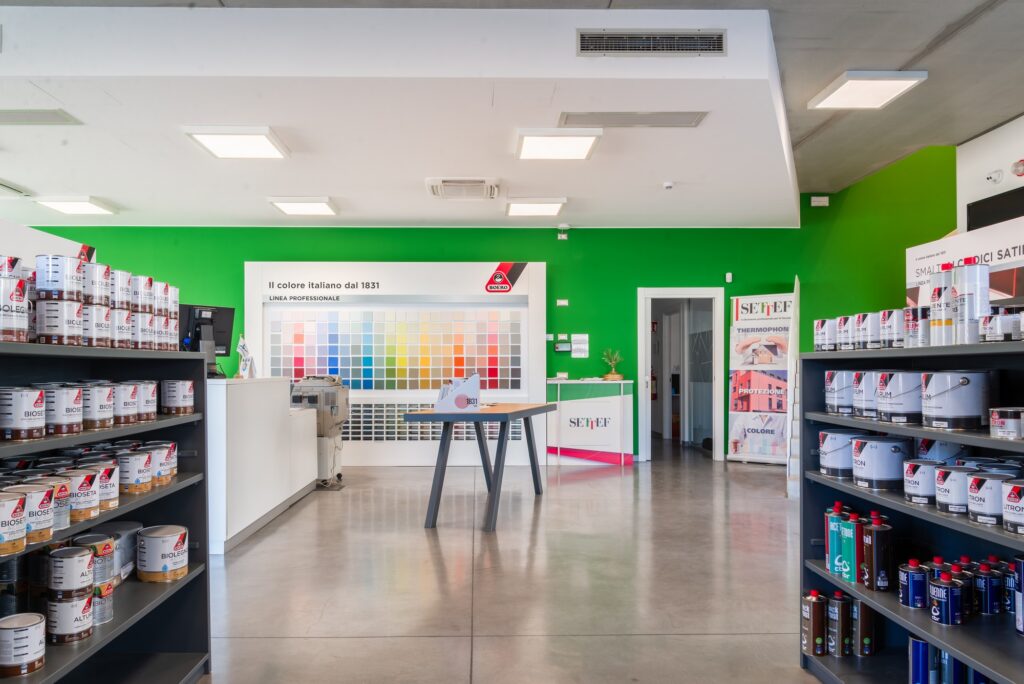 Interno del negozio Colorificio Bergamini a Bolzano con scaffali di prodotti vernicianti, banco accoglienza e parete campionatura colori.